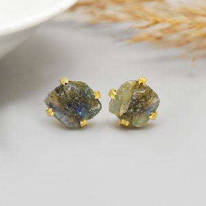 Pode incluir: Um par de brincos de pressão de labradorite. Cada brinco apresenta uma pedra bruta multicolorida com tons de azul, verde e dourado, engastada em metal dourado. Os brincos são exibidos em uma superfície branca.