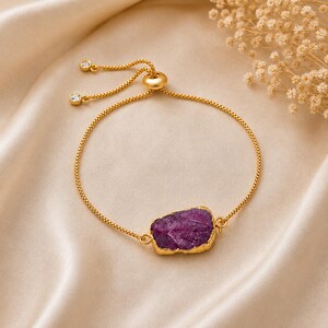 May include: A gold-toned adjustable bracelet featuring a central, irregular, deep purple druzy stone with a textured surface. The bracelet has a thin box chain, a sliding bead mechanism for sizing, and two small clear stones at the ends of the chain.