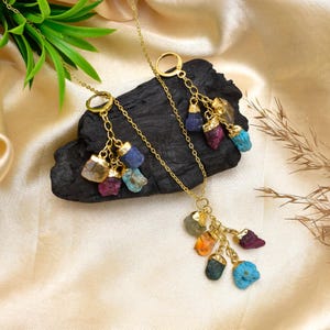 May include: A gold-colored necklace and earrings set featuring raw, colorful gemstones. The necklace and earrings have a delicate chain design, with each piece adorned with a cluster of various-colored stones, including blue, red, and yellow. The jewelry is displayed on a dark, textured surface.