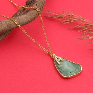 Könnte beinhalten: Eine goldfarbene Halskette mit Anhänger. Der Anhänger ist ein hellgrüner, unregelmäßig geformter Stein mit goldfarbenem Rand. Der Buchstabe "A" ist mit kleinen, klaren Kristallen besetzt und am Stein befestigt.