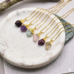 Peut inclure: Un ensemble de six colliers à chaîne dorée, chacun doté d'un pendentif en pierre brute non taillée. Les pierres incluent des teintes violet foncé, violet clair, vert pâle, rose tendre, rouge profond et jaune translucide, serties dans des capuchons dorés.