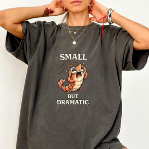 May include: Dark gray t-shirt with the text "SMALL BUT DRAMATIC" above a cartoon shrimp illustration. The shrimp is orange and white with a distressed expression. The shirt has short sleeves and a relaxed fit.
