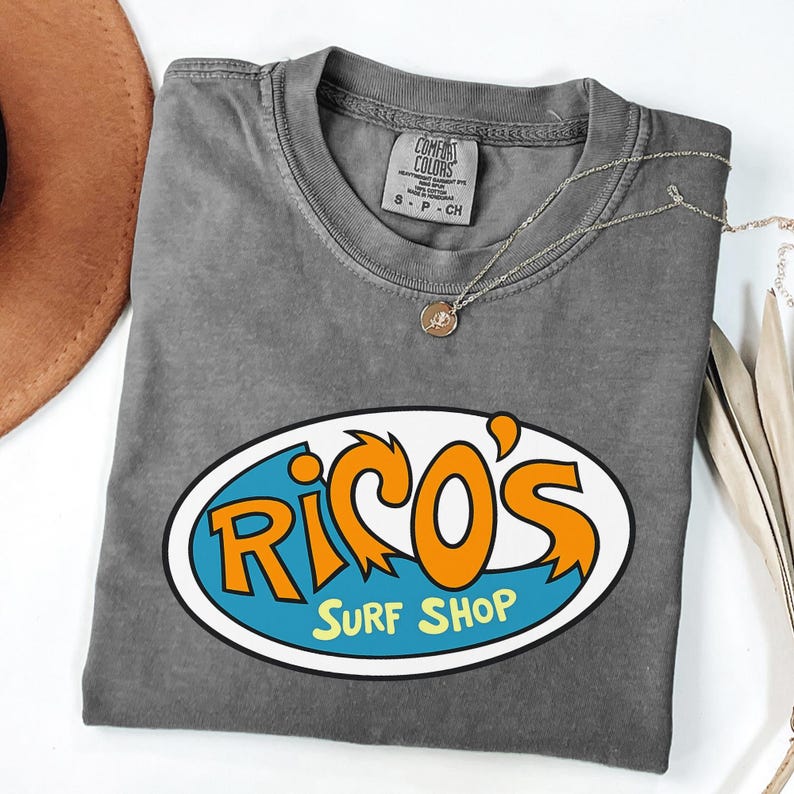 May include: A grey crew neck t-shirt with a colourful graphic of "Rico's Surf Shop" in orange and blue. A silver necklace with a small pendant is draped across the shirt.