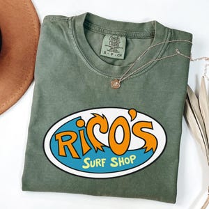 May include: An olive green t-shirt featuring the text "Rico's Surf Shop" in an oval design. The text is in orange and blue. A gold necklace with a pendant is on the shirt. The shirt is made of a soft material.