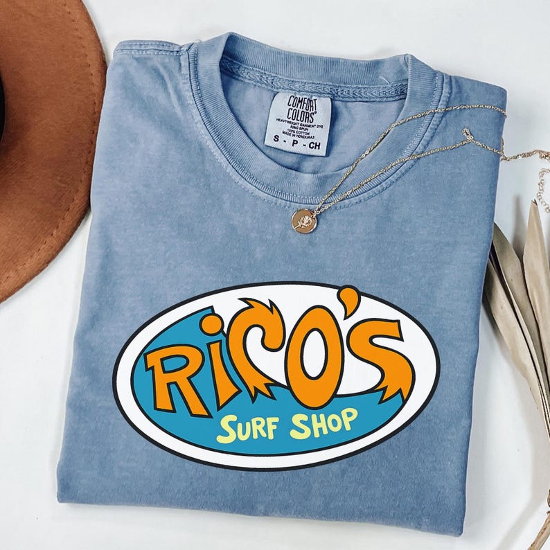 May include: A blue t-shirt featuring the text "Rico's Surf Shop" in orange and white, within a blue and white oval. The shirt has a crew neck and short sleeves. A gold necklace with a pendant is draped across the shirt.