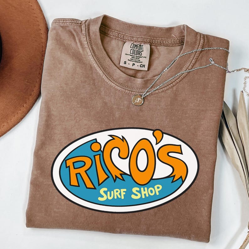 May include: A brown t-shirt featuring the text "Rico's Surf Shop" in orange and blue within an oval design. The shirt has a crew neck and short sleeves. A silver necklace with a small pendant is draped across the shirt.
