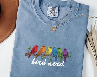 May include: A light blue t-shirt with a graphic of six colorful birds perched on a branch, with the words "bird nerd" in white script below. A gold necklace with a pendant is draped across the shirt.