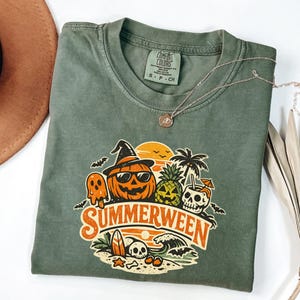 May include: An olive green crew neck t-shirt featuring a graphic print with the text "SUMMERWEEN". The design includes a jack-o'-lantern wearing sunglasses and a witch hat, a pineapple, a skull in a drink, a surfboard, and a beach scene.