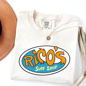 May include: Cream-coloured t-shirt featuring the text "Rico's Surf Shop" in orange and blue. The shirt is folded, with a gold necklace and a brown hat nearby. The shirt is made of a soft material.