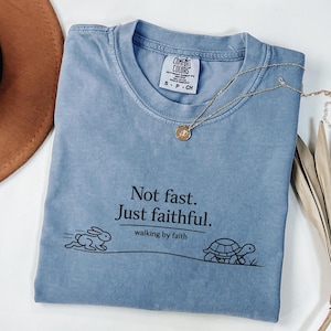 May include: A light blue t-shirt with the text "Not fast. Just faithful. walking by faith." A gold necklace with a pendant is draped across the shirt. A drawing of a rabbit and a turtle are below the text.