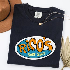 May include: A navy blue t-shirt featuring an oval graphic with the words "Rico's Surf Shop" in orange and white on a blue background. A gold necklace and a brown hat are also visible.