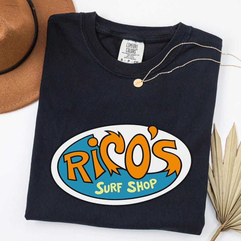 May include: A navy blue t-shirt featuring an oval graphic with the words "Rico's Surf Shop" in orange and white on a blue background. A gold necklace and a brown hat are also visible.