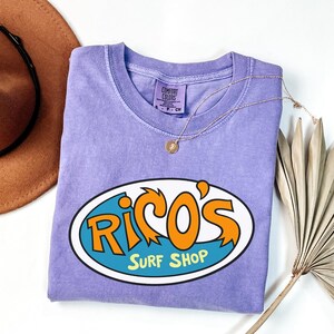 May include: A lavender Comfort Colors t-shirt with the text "Rico's Surf Shop" in orange and blue. A gold necklace with a pendant rests on the shirt. A brown hat and dried plant are in the background.