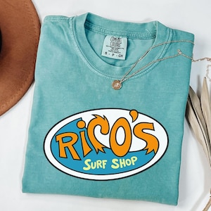 May include: A turquoise Comfort Colors t-shirt featuring the text "Rico's Surf Shop" in an oval design. The text is orange and white with a blue background. A gold necklace with a pendant is draped across the shirt.