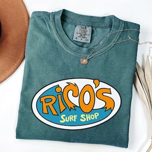 May include: A teal Comfort Colors t-shirt featuring the text "Rico's Surf Shop" in orange and white on a blue oval. A gold necklace with a pendant is draped across the shirt. The shirt is folded.