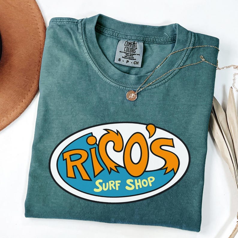 May include: A teal Comfort Colors t-shirt featuring the text "Rico's Surf Shop" in orange and white on a blue oval. A gold necklace with a pendant is draped across the shirt. The shirt is folded.