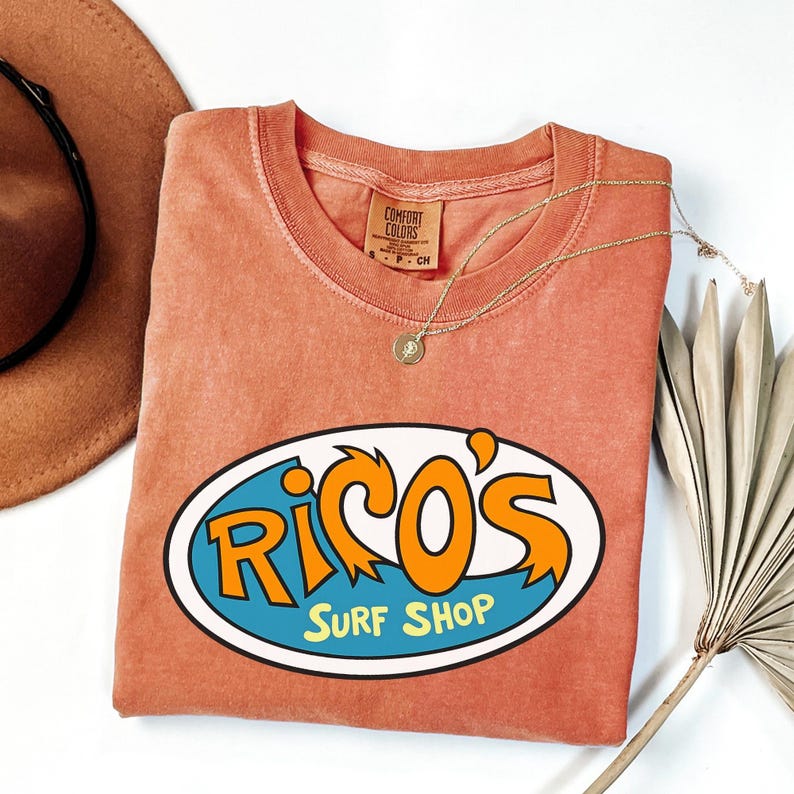 May include: A coral-coloured t-shirt with the text "Rico's Surf Shop" in a blue and orange oval design. A gold necklace and a brown hat are next to the shirt. A dried palm leaf is also in the image.