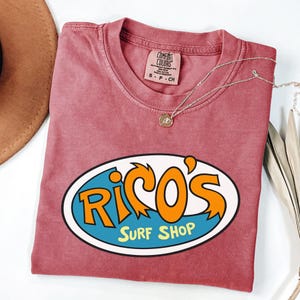 May include: A faded red t-shirt with a crew neck and the text "Rico's Surf Shop" in an oval design. The design features orange and blue lettering. A silver necklace with a small pendant is on the shirt.