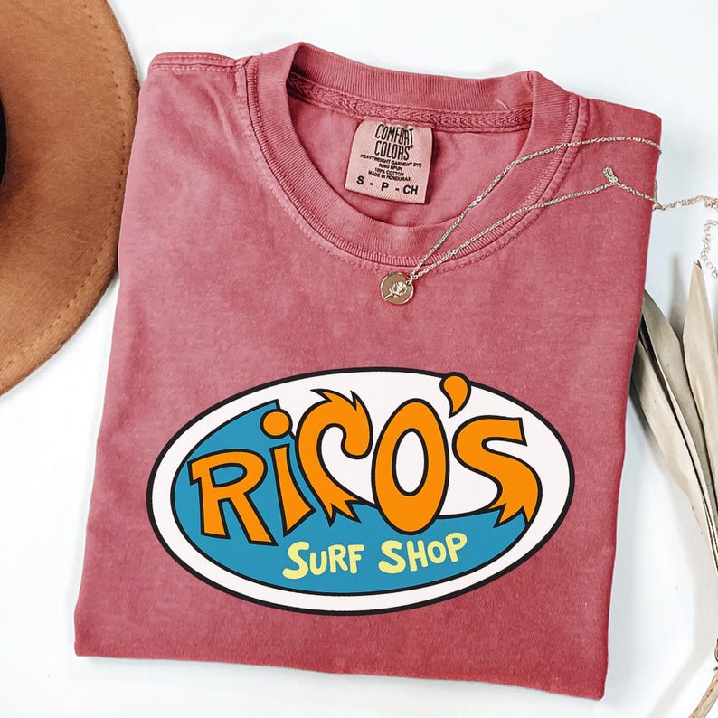 May include: A faded red t-shirt with a crew neck and the text "Rico's Surf Shop" in an oval design. The design features orange and blue lettering. A silver necklace with a small pendant is on the shirt.
