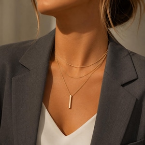 Peut inclure: Un ensemble de colliers dorés superposés composé de trois chaînes distinctes. La couche supérieure est une chaîne fine et lisse, la couche intermédiaire est une chaîne à maillons délicats, et la couche inférieure porte un pendentif barre rectangulaire.