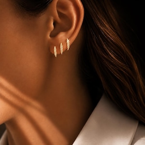 Op de afbeelding: Drie goudkleurige oorbellen met een lineair ontwerp, elk bezet met kleine, sprankelende steentjes. De oorbellen worden op het oor gedragen, met de nadruk op het ontwerp en de plaatsing van de sieraden. De oorbellen zijn een soort sieraad.