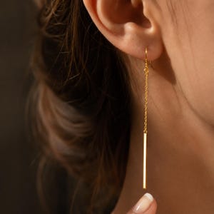 Peut inclure: Une boucle d'oreille pendante dorée composée d'une fine chaîne délicate suspendue à un crochet, se terminant par une tige rectangulaire polie et élancée qui pend verticalement le long du cou, créant une silhouette épurée et allongée.