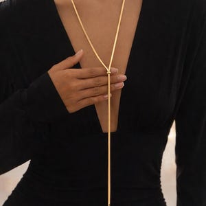 Peut inclure: Un collier lariat doré avec une chaîne délicate. Le collier présente un design simple avec un détail de nœud et une longue goutte fine. Le collier est porté sur une robe noire.