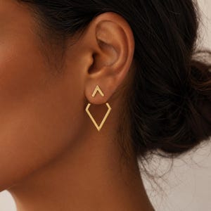 Puede incluir: Un par de pendientes dorados. Los pendientes presentan un diseño geométrico, con una pequeña forma de V invertida sobre una forma de diamante más grande. Los pendientes se llevan en una oreja, enfocando la joyería.