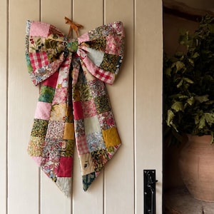 May include: A large decorative bow constructed from a patchwork of various fabric squares featuring floral, gingham, and solid patterns in shades of pink, green, yellow, and red. The bow is suspended by a thin brown ribbon loop at the top center.