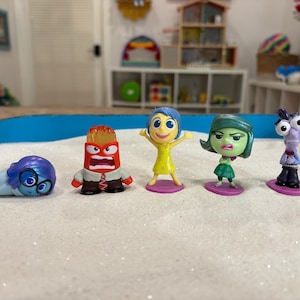 May include: Five colorful figurines from the animated movie "Inside Out" are arranged on a white, glittery surface within a blue tray. The characters include Sadness, Anger, Joy, Disgust, and Fear, each with distinct features and expressions.