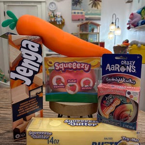 May include: A Jenga game box, a Squeeezy peach toy, a Crazy Aaron's Goodnight Cactus thinking putty tin, and a Squeeeze Butter toy box arranged with a large orange carrot-shaped toy resting on top of the items in a colorful display.