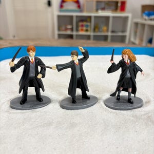 May include: Three miniature figurines of characters from the Harry Potter series. Each figure is wearing a black robe, a gray vest, and a white shirt with a red and yellow tie. They are holding wands and standing on gray bases.