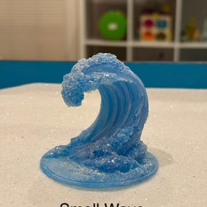 May include: A light blue wave sculpture with a textured surface and a base. The sculpture has a frosted appearance, with the text "Small Wave" printed in black at the bottom. The wave is a decorative item.