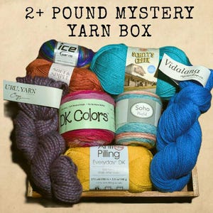 May include: A wooden tray filled with various colorful yarn skeins. The image includes skeins of blue, teal, orange, purple, pink, and yellow yarn. Text on the image reads "2+ POUND MYSTERY YARN BOX". Labels on the yarn include "ice yarns", "DK Colors", and "Vidalana".