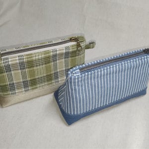 May include: Two rectangular pencil cases. One is green plaid with a beige base and a bronze zipper. The other is blue and white striped with a denim base and a bronze zipper. Both cases have a small fabric loop attached to the zipper.