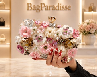 May include: A rectangular clutch bag featuring a cream-coloured base adorned with an array of three-dimensional fabric flowers in shades of pink, white, and gold, accented with clear beads, sequins, and a gold-toned metal clasp at the top.