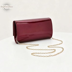 Puede incluir: Un clutch de charol burdeos con una cadena dorada. El bolso rectangular tiene un acabado brillante y un cierre de solapa. La marca "BagParadisee" está en la esquina superior izquierda.