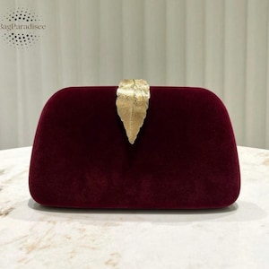 Puede incluir: Bolso de mano de terciopelo burdeos con cierre dorado en forma de hoja. El bolso rectangular tiene bordes redondeados y un aspecto suave y texturizado. La marca "BagParadisee" está en la esquina superior izquierda.
