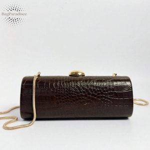 Può includere: Una borsa a mano cilindrica marrone con una texture a pelle di coccodrillo. La borsa ha una chiusura color oro e una tracolla a catena dorata. Il nome del marchio "BagParadisee" è stampato in alto a sinistra.