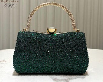 Puede incluir: Un bolso de mano verde con cuentas, con un asa y un cierre dorados. El clutch rectangular presenta una superficie brillante y texturizada. El asa es arqueada e incrustada con pequeñas piedras brillantes. El bolso está sobre un fondo neutro.