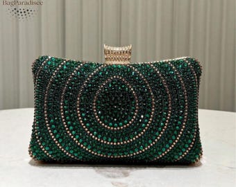 Puede incluir: Clutch verde esmeralda con cuentas y cierre dorado. El bolso rectangular presenta círculos concéntricos de pequeñas cuentas brillantes. El cierre está adornado con pequeños cristales transparentes. Un accesorio formal.