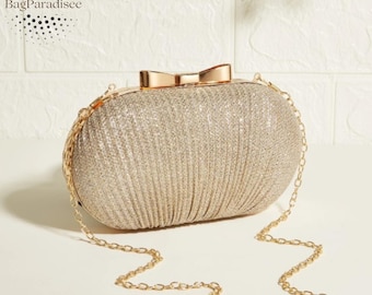 May include: A gold glitter clutch with a bow closure and a gold chain strap. The oval-shaped evening bag has a textured, pleated design. The text "BagParadisee" is in the upper left corner.