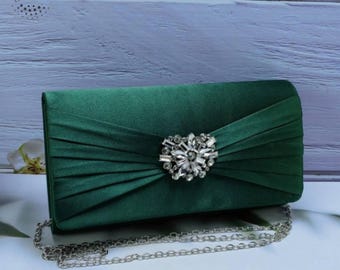 Puede incluir: Bolso de mano de satén verde oscuro con una correa de cadena plateada. La parte delantera del bolso presenta un adorno decorativo de plata y cristal brillante. El bolso de mano es rectangular con la parte superior curvada.