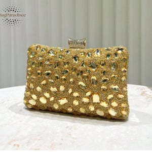 Puede incluir: Un bolso de mano dorado con forma rectangular. La superficie está cubierta de pequeñas cuentas redondas y piedras preciosas transparentes más grandes, de forma irregular. Un pequeño cierre rectangular con piedras transparentes en la parte superior. La marca "BagParadisee" está en la esquina superior izquierda.