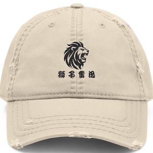 以下が含まれることがあります： 黒いライオンの頭のグラフィックと日本語のテキストが特徴の、使い古されたベージュの野球帽。帽子は湾曲したつばと、使い古された外観をしています。帽子には上部のボタンと調節可能なストラップが付いています。