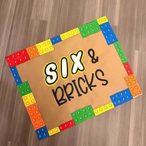 May include: A rectangular sign with a brown background and colorful block-style borders. The sign features the text "SIX & BRICKS" in a playful font, with the word "SIX" in white with a black outline and "BRICKS" in black.