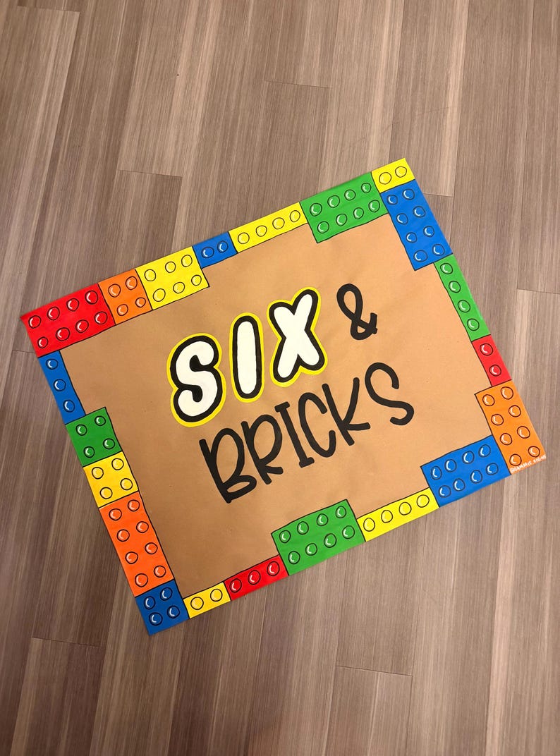 May include: A rectangular sign with a brown background and colorful block-style borders. The sign features the text "SIX & BRICKS" in a playful font, with the word "SIX" in white with a black outline and "BRICKS" in black.