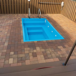 Könnte beinhalten: Ein kleines, rechteckiges, blaues Schwimmbecken mit Stufen ist in eine Backstein-Terrasse eingelassen. Das Becken hat Metallgeländer. Die Terrasse ist von einem Holzzaun und einer braunen Terrasse umgeben.