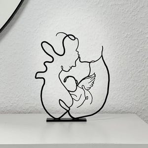 Könnte beinhalten: Schwarze Metallskulptur, die eine Mutter und ein Kind in einer durchgehenden Linie darstellt. Die Skulptur zeigt eine Mutterfigur, die ein Baby mit Engelsflügeln umarmt. Das Kunstwerk ist auf einem kleinen schwarzen Sockel montiert.