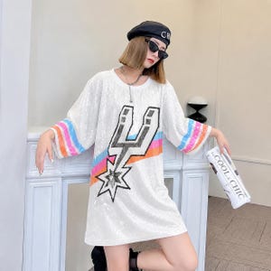 May include: White sequined dress with a graphic design featuring a star and lightning bolt. The sleeves have rainbow stripes. The dress is knee-length and has a loose fit. The model is wearing a black beret and sunglasses.